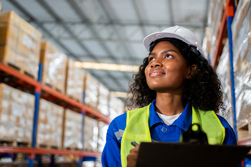 Happy Professional woman Worker Wearing Safety Vest and Hard Hat Smiling on Camera. In the Background Big Warehouse with Shelves full of Delivery Goods.