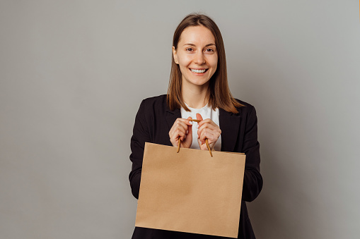 Wide smiling woman is holding a blank kraft shopping bag with copy space over grey background.