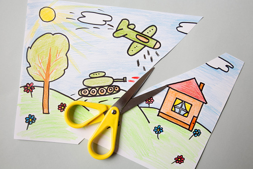Children's drawing about the war with a tank and a plane flying over the house in the field. Scissors cut a piece of paper with a children's drawing about war and peace.