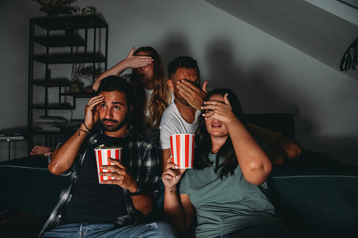 A group of four friends are watching TV at home while they are eating popcorn. They are having fun together.