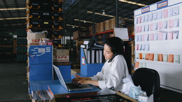 Businesswoman working on laptop computer and talking on mobile phone at Automobile Industry warehouse