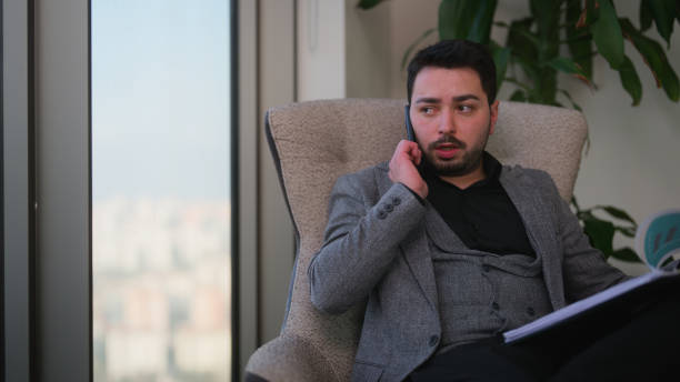 istock Young businessman standing next to window and talking on phone while checking papers and documents in modern office 1471699039