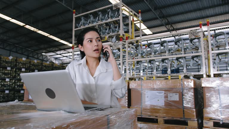 Businesswoman working on laptop computer and talking on mobile phone at Automobile Industry warehouse