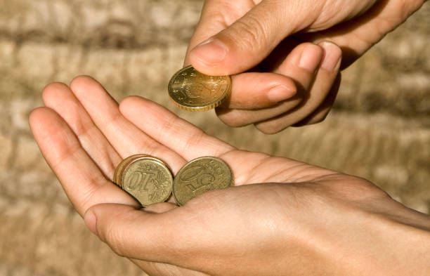 A human hand pouring coins into a wooden piggy bank placed on a bright turquoise background. The shot symbolizes savings, financial planning, and accumulation of funds.