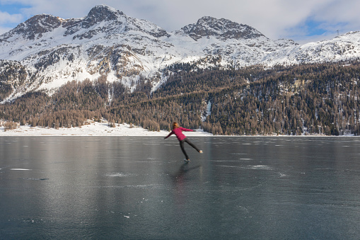 Subject: Horizontal view of a teenage girl in a figure skating pose stretching across an outdoor winter pond, with the bright afternoon sun shining on the mountains of Piz San Gian, Piz Surlej, Munt Arlas, casting a long shadow on his figure as he glides on one foot on the smooth ice. Lake Silvaplana,  canton of Graubunden, Engadine, Switzerland