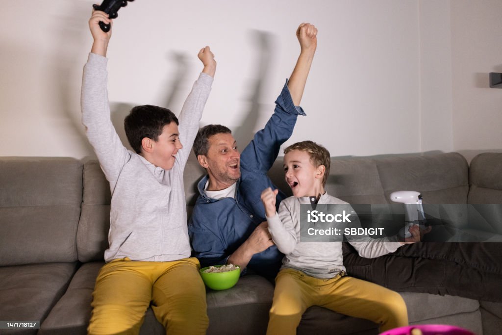 Father with sons celebrate their win at the video game on PlayStation Caucasian family, father and sons having fun while playing video games on PlayStation Father Stock Photo