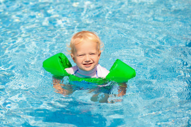 Baby with inflatable armbands in swimming pool. Little boy learning to swim in outdoor pool of tropical resort. Swimming with kids. Healthy sport activity for children. Sun protection. Swim aids.