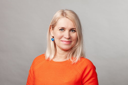 Close-up studio portrait of a 50 year old blonde woman with long hair in an orange sweater against a gray backgroud