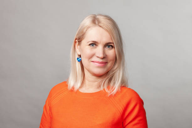 Close-up studio portrait of a 50 year old blonde woman with long hair in an orange sweater against a gray backgroud stock photo