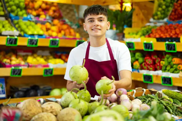 Young salesman working and posing in store near counter with fresh round bottlegourd vegetable