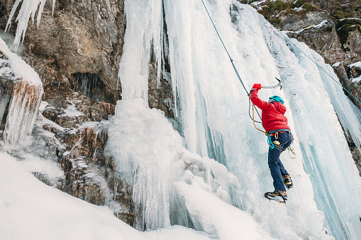 Ice climber dressed in warm winter climbing clothes, safety harness and helmet climbing frozen waterfall using two Ice climbing axes and crampons. Active people and sports activities concept image.