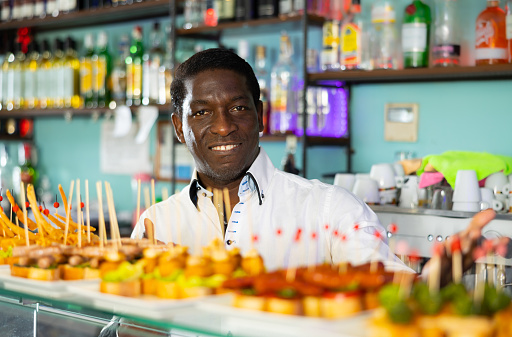 African american male bartender standing behind bar counter in pub and offering appetizing plate of pinchos