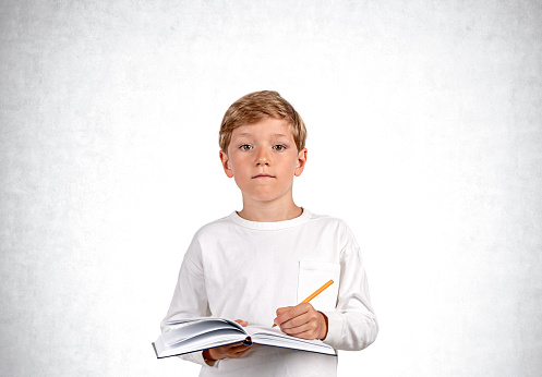 Pondering handsome boy in casual wear standing holding notebook and taking notes near empty white wall in background. Concept of inspired kid, education, learning, studying, student