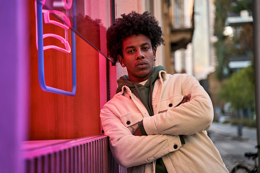 Confident cool African American hipster guy standing at city street near neon light sign. Rebel gen z male model looking at camera outside advertising young generation culture life entertainment.