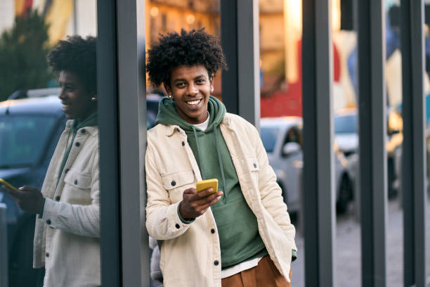 улыбается молодой афроамериканский парень, пользующийся мобильным телефоном, стоящий на городской улице. - gen z стоковые фото и изображения