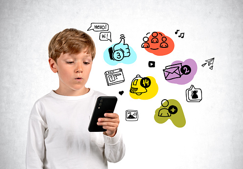 Pensive boy looking at phone in hand, colorful social media doodle icons on grey concrete background. Concept of online education and communication
