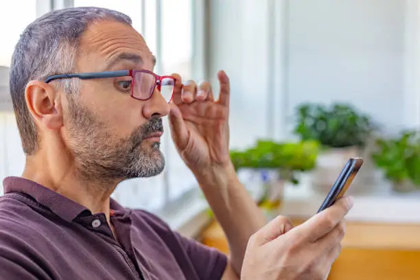 Spain. Adult man with beard putting on presbyopic glasses to see closely the mobile phone screen