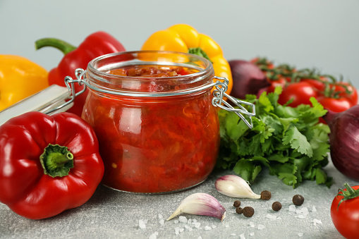 Glass jar of delicious canned lecho and fresh ingredients on light grey table, closeup