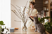 A happy girl hangs Easter eggs on a willow bouquet at home with a little kitten. Funny pets. Easter decoration