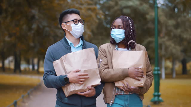 Diverse couple in safety mask walk outdoors with grocery bags. Realtime