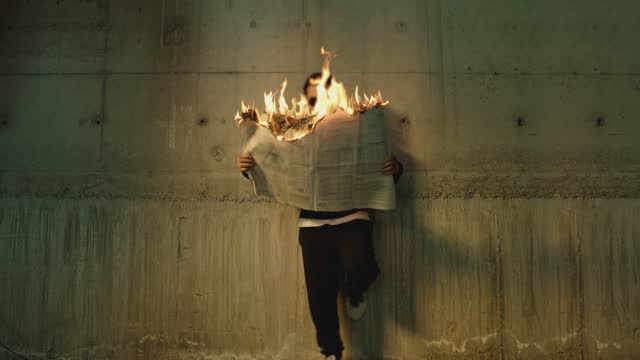 Footage of male artist with sunglasses standing near wall , holding and reading newspaper during it burns . Concept or symbol of fake or forbidden news   . Man reads burning magazine . Slow motion