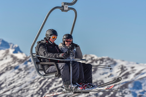 Two caucasian skiers in ski clothing in a chair lift going up with a snowy mountain in the background, 29 december 2016