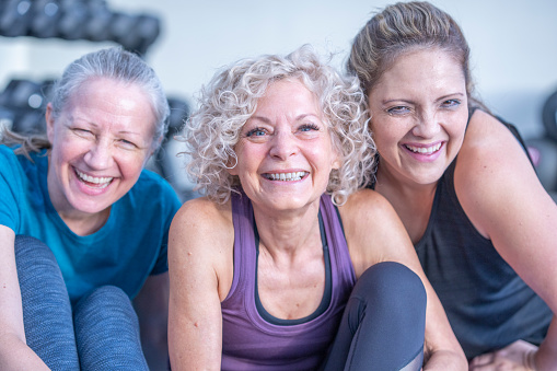 A small group of three mature women huddle in closely together on the floor in a gym as they pose for a portrait together after class. 
 They are each dressed comfortably in athletic wear and are smiling as they and laughing.
