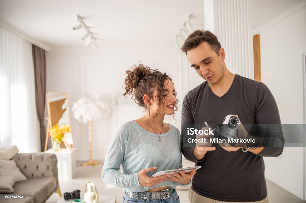 A young couple, a man and a woman, are setting up a security camera for home surveillance in their apartment A young couple installs a security camera in their new home, an alarm system, they plan where best to install the video surveillance of their new apartmentv Domestic Life Stock Photo