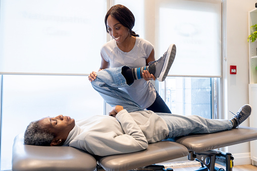 A young female Chiropractor of African decent, works with a senior woman who is laying prone on her treatment table as she evaluates the problem.  She is moving the patients leg in various positions and stretches to help pinpoint the trouble area and adjust it accordingly.