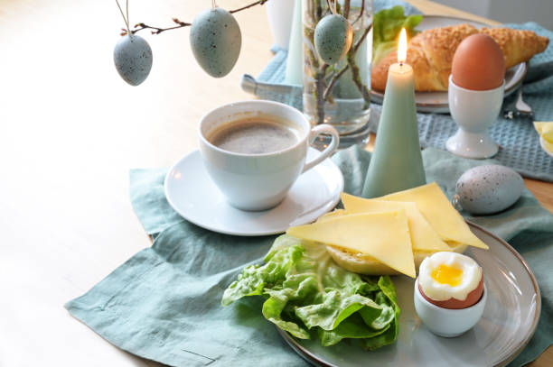 istock Breakfast table setting with bread roll, cheese, boiled egg and coffee on a pastel turquoise napkin, hanging Easter eggs and candle as decoration, copy space, selected focus 1471751812