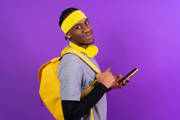 Black ethnic man with backpack and yellow headphones on a purple background, student concept