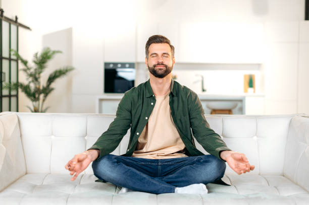 soddisfatto calmo felice uomo barbuto caucasico, in abiti casual, siede su un divano in posizione di loto con gli occhi chiusi in salotto, rilassandosi dopo una giornata di lavoro, meditando, sorridendo - spirituality yoga zen like meditating foto e immagini stock