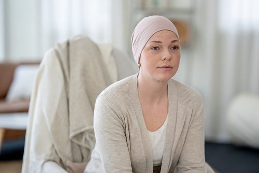 A young woman battling cancer sits in the comfort of her own home between treatments as she poses for a portrait.  She is dressed comfortably and wearing a headscarf to keep warm.