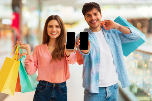Cheerful Couple Showing Smartphone Screen Recommending Mobile Shopping Application Posing With Colorful Shopper Bags In Modern Hypermarket. Selective Focus On Phones. Mockup