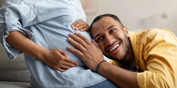 Photo of Black Husband Listening To Pregnant Wife's Belly Indoor, Panorama, Cropped