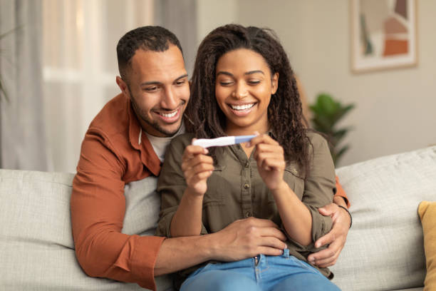 happy african american couple holding positive pregnancy test and smiling, man hugging woman on sofa at home - domestic life young family family child imagens e fotografias de stock