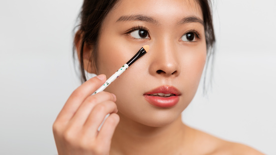 Natural Make Up. Pretty young Asian lady using cosmetic brush to apply concealer under her eyes, white studio background, closeup headshot banner. Teenage girl putting on makeup for circles correction