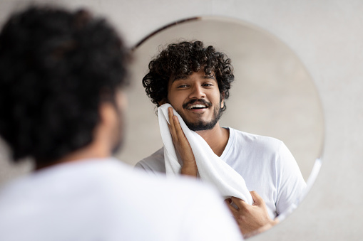 Everyday routine concept. Excited indian man wiping his face with towel while standing in modern bathroom, looking at reflection in the mirror. Male facial skincare routine concept