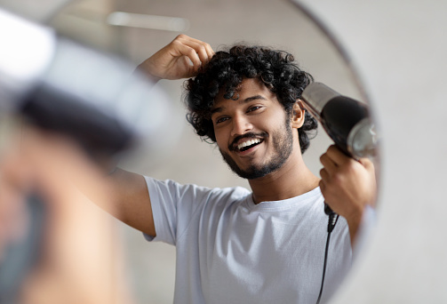 Excited indian man using hairdryer after shower, drying his curly hair, making hairdo, taking care of himself in the morning, smiling at his reflection in mirror. Everyday hygiene concept