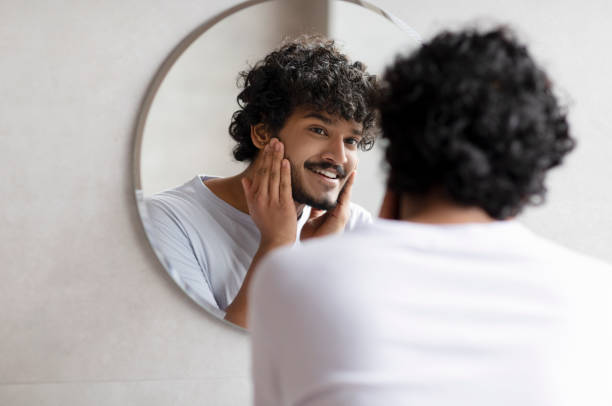 istock Attractive bearded indian man touching face applying moisturizer on face, standing near mirror in modern bathroom 1471775598