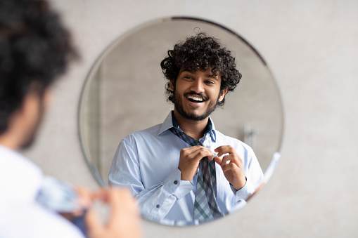 Happy indian businessman putting on necktie while looking in the mirror, standing in modern bathroom, free space