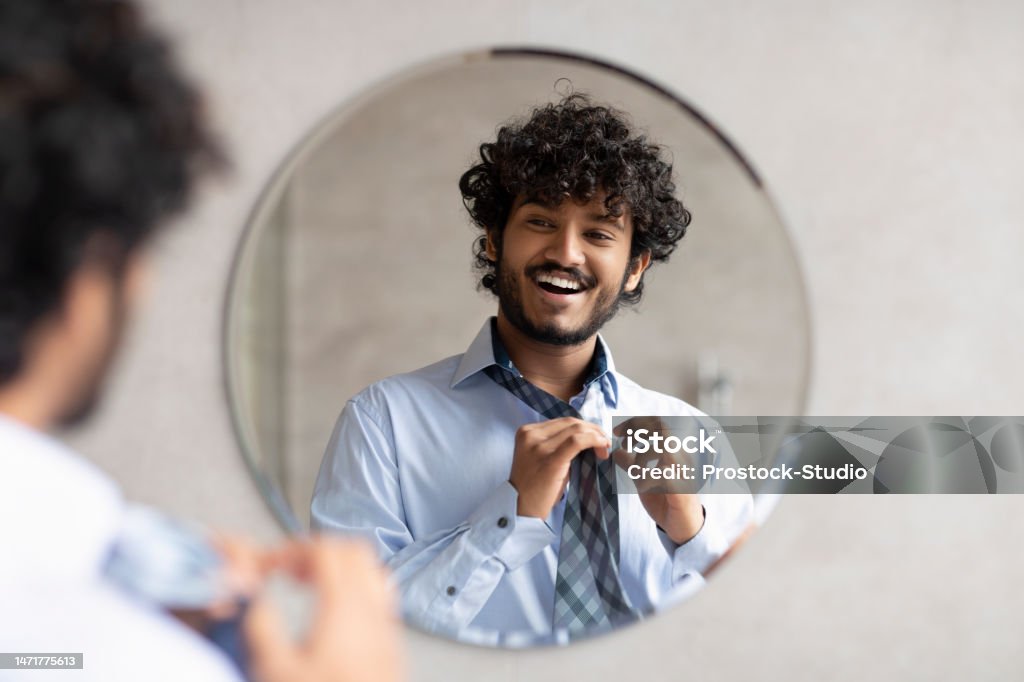 Happy indian businessman putting on necktie while looking in the mirror, standing in modern bathroom, free space Happy indian businessman putting on necktie while looking in the mirror and smiling at his reflection, standing in modern bathroom interior, free space Necktie Stock Photo