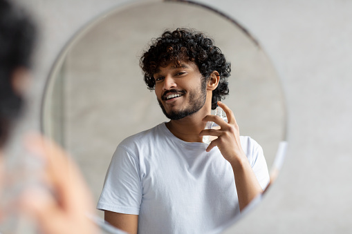 Happy indian man looking in mirror spraying on cologne in bathroom interior, doing morning skincare routine. Beauty skin care, moisturizers products and perfume