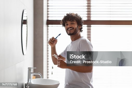 istock Oral care concept. Happy bearded indian guy in white t-shirt brushing teeth with paste, smiling at camera in bathroom 1471775768