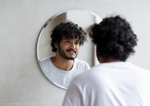 Handsome indian guy looking at his reflection in the mirror, standing in modern bathroom at home, doing morning beauty routine. Self care concept