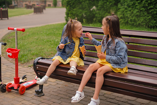 Sisters share ice cream with different flavors with each other. The girl gives a dessert from a spoon to a friend to try. The children sat down to rest on a bench after riding scooters.