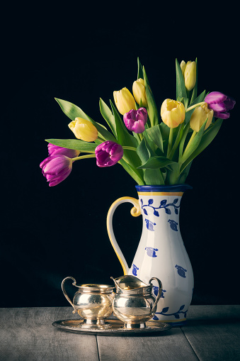 A still life image of a pitcher filled with yellow and purple tulips with sterling silver pieces.