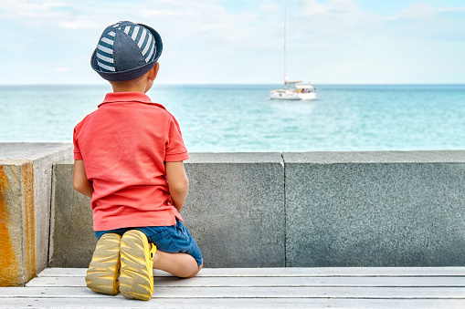 Dreaming little boy with panama looks at yacht sailing on sea from bench on seafront backside view. Toddler kid rests on city embankment