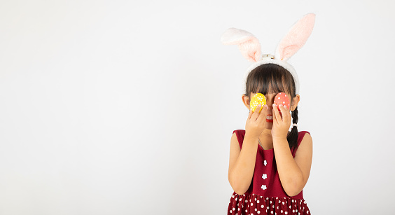 Happy Easter Day. Smile Asian little girl wearing easter bunny ears holding colorfull eggs closes eyes with testicles isolated on white background with copy space, Happy child in holiday