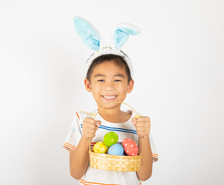 Happy Easter Day. Smile Asian little boy wearing easter bunny ears holding basket of full colorful eggs smiles broadly isolated on white background with copy space, Happy child in holiday